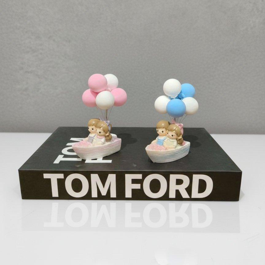 Two small figurines with balloons on a black platform with 'Tom Ford' branding.