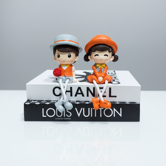 Two small figurines on a Chanel and Louis Vuitton book with a gray background