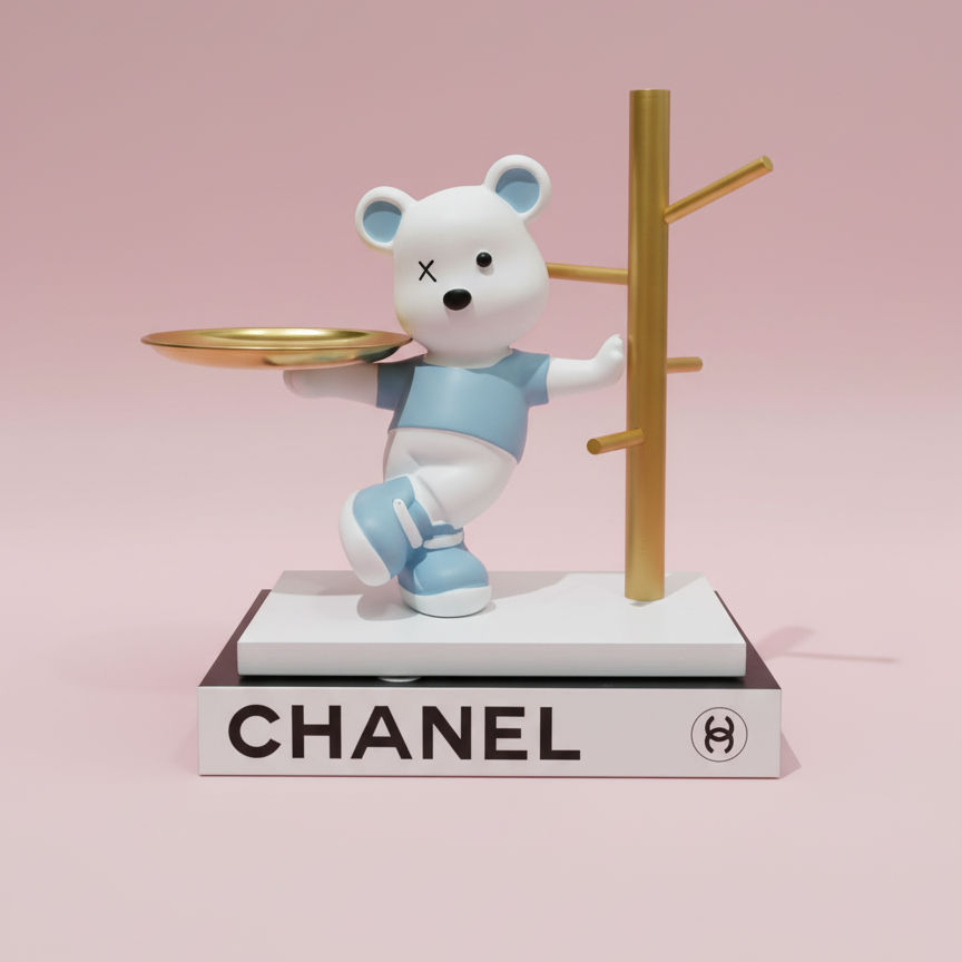 White teddy bear figurine with blue outfit holding a tray on a Chanel book with pink background