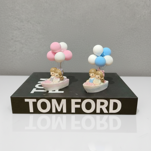 Two small figurines with balloons on a black platform with 'Tom Ford' branding.