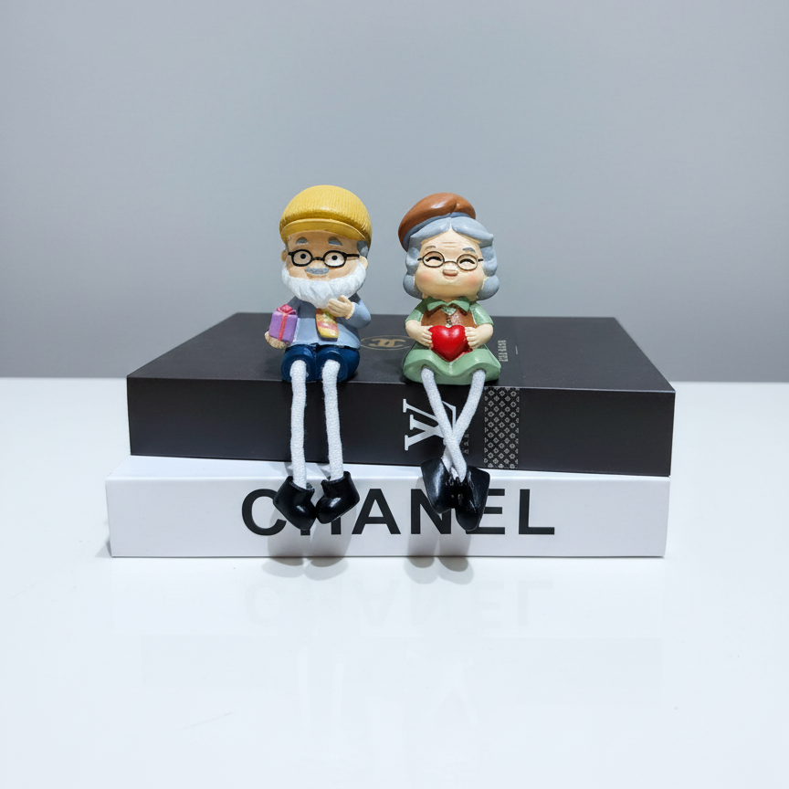 Two figurines of an older couple sitting on a Chanel box with a gray background